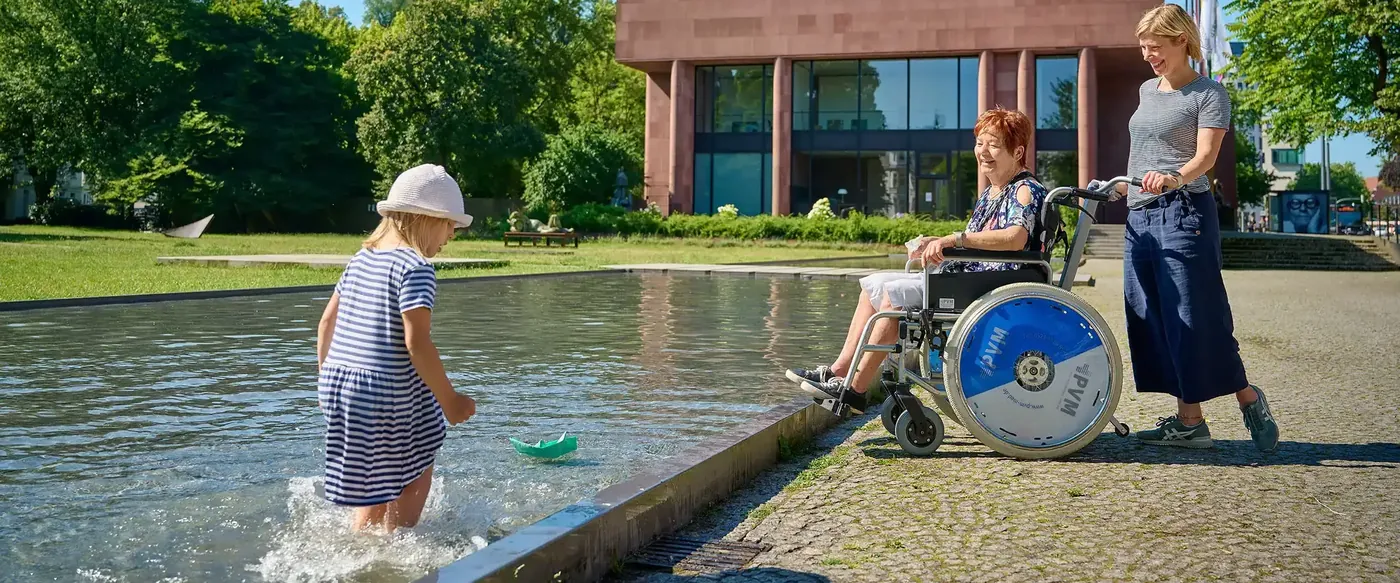 Vor der Kunsthalle Bielefeld: eine Frau im Rollstuhl mit Begleiterin am Wasserbecken, ein Kind spielt mit einem Papierboot