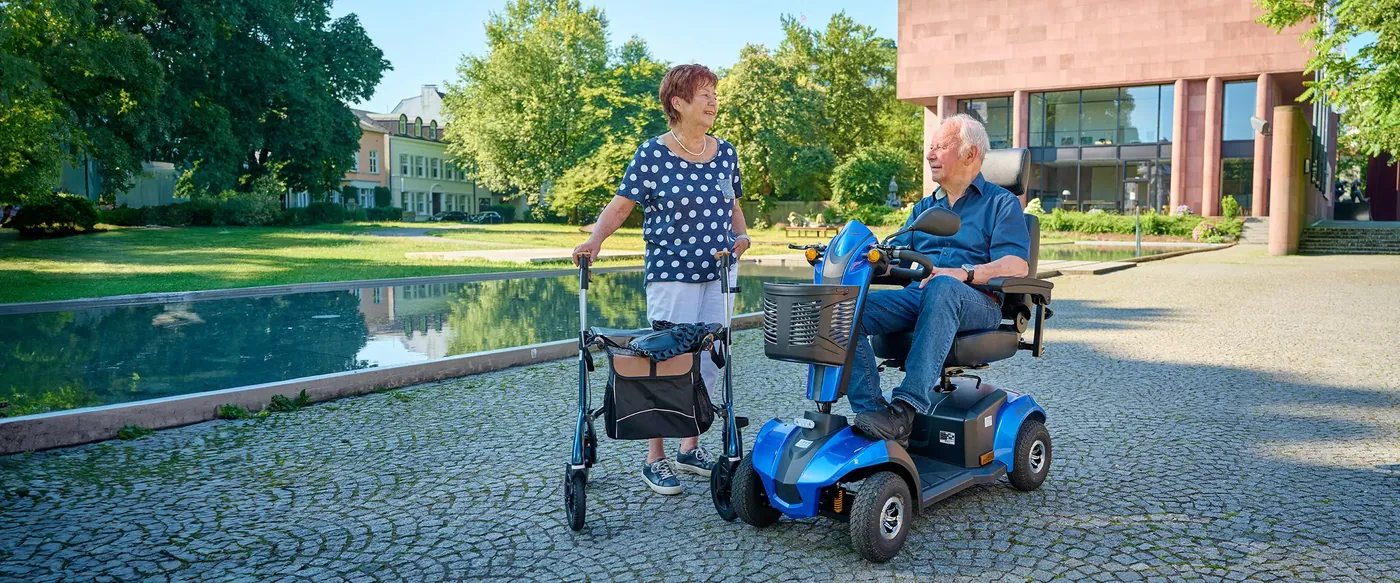 Kundin mit Rollator und Kunde auf einem Elektromobil im Park